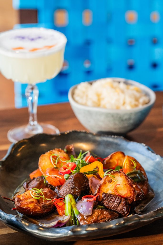 Plate of glazed grilled meat with peppers and herbs on a dark dish with a blue background and bowl of rice in the background