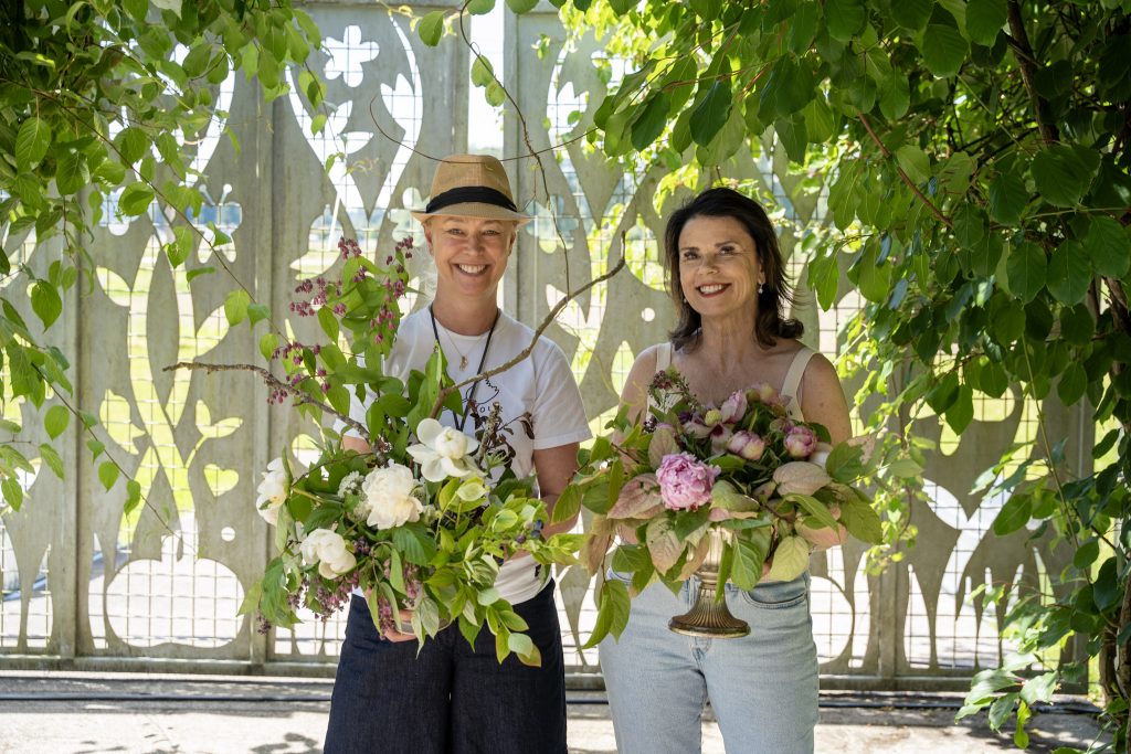 Two women smile while holding large bouquets of flowers among green leaves and a decorative fence behind them outdoors